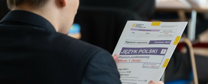 Uczeń w garniturze trzyma arkusz egzaminu maturalnego z języka polskiego.