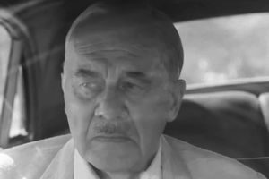 Mężczyzna w jasnym garniturze siedzi w samochodzie, ma poważny wyraz twarzy.
