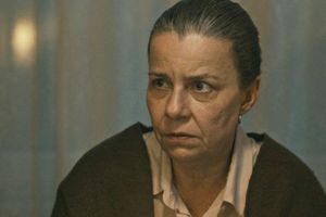 Kobieta z wyraźnym przejawem zmartwienia, siedzi przy stole z tłem zasłoniętym firanką.