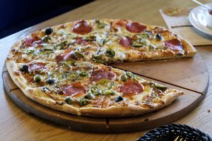Pizza z pepperoni, oliwkami i papryką, część już pokrojona, podana na drewnianym talerzu.