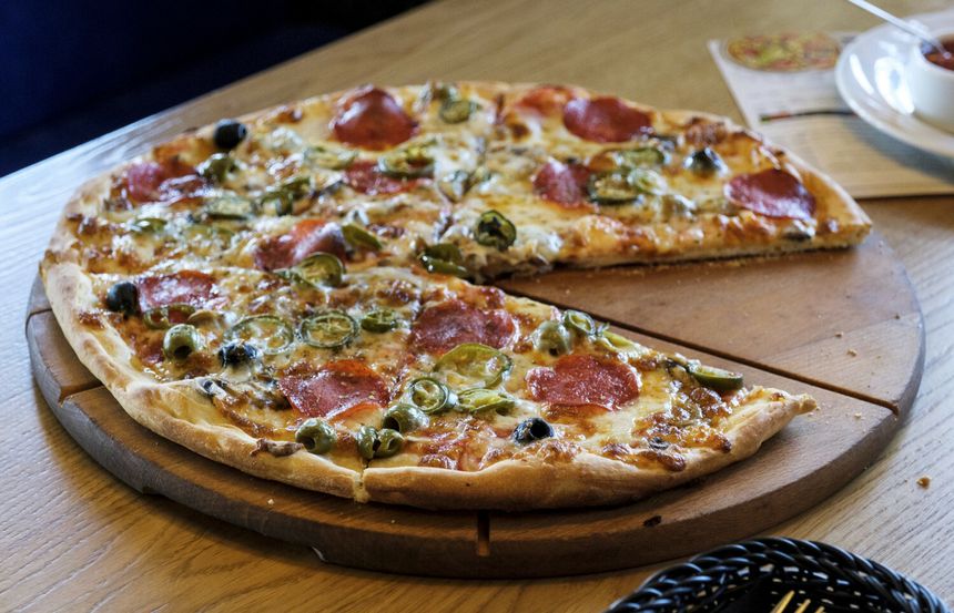 Pizza z pepperoni, oliwkami i papryką, część już pokrojona, podana na drewnianym talerzu.