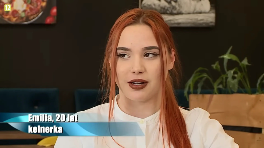 Młoda kobieta z rudymi włosami w białej koszuli, mówi w kawiarnianym otoczeniu.