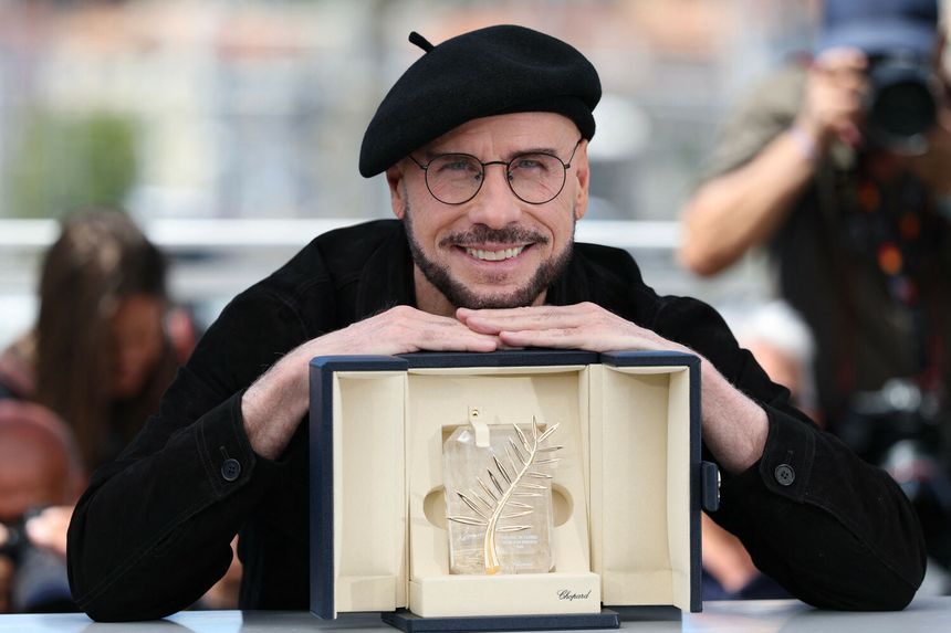 Mężczyzna z okularami i beretem uśmiecha się, trzymając trofeum na tle z tłumem.