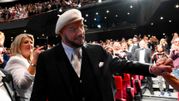 Mężczyzna w białym kapeluszu wita się z publicznością w tle stoją ludzie w formalnym ubiorze.
