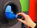 Osoba wrzuca niebieską butelkę do otworu recyklingowego maszyny. Tło w różnych kolorach.