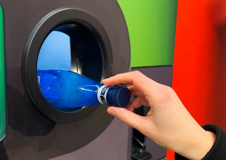 Osoba wrzuca niebieską butelkę do otworu recyklingowego maszyny. Tło w różnych kolorach.