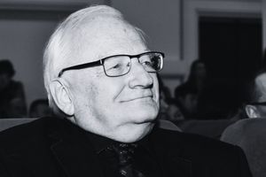 Mężczyzna w okularach, uśmiechający się, siedzi wśród publiczności w sali.