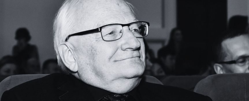 Mężczyzna w okularach, uśmiechający się, siedzi wśród publiczności w sali.