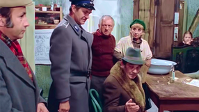 Grupa osób w wiejskiej chatce, przy stole, w scenie z filmu komediowego.