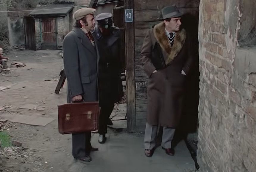 Trzech mężczyzn w eleganckich strojach stoi w zniszczonym otoczeniu. Na zdjęciu widoczny jest sierżant policji.