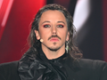 Michał Szpak z długimi włosami, dramatycznym makijażem, w ciemnym stroju, na tle czerwono-czarnym.