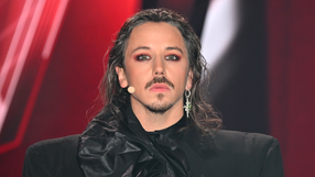 Michał Szpak z długimi włosami, dramatycznym makijażem, w ciemnym stroju, na tle czerwono-czarnym.