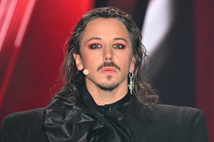 Michał Szpak z długimi włosami, dramatycznym makijażem, w ciemnym stroju, na tle czerwono-czarnym.