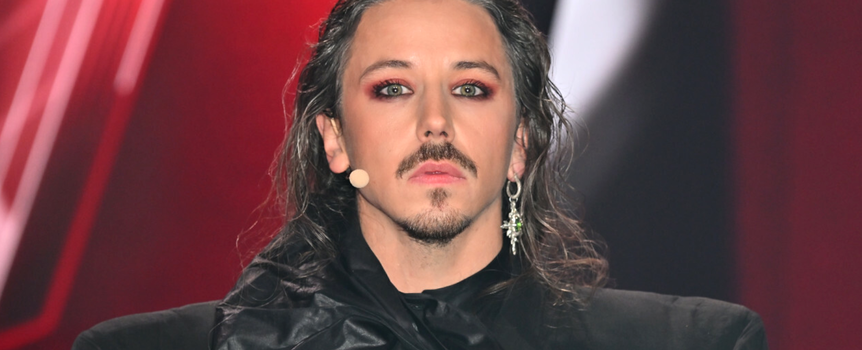 Michał Szpak z długimi włosami, dramatycznym makijażem, w ciemnym stroju, na tle czerwono-czarnym.