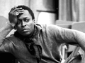 Miles Davis w szarym ubraniu z papierosem, zamyślony, siedzi na krześle w pomieszczeniu.