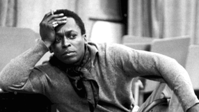 Miles Davis w szarym ubraniu z papierosem, zamyślony, siedzi na krześle w pomieszczeniu.