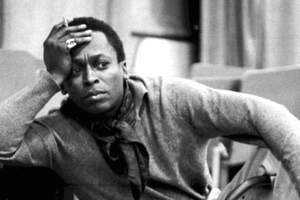Miles Davis w szarym ubraniu z papierosem, zamyślony, siedzi na krześle w pomieszczeniu.