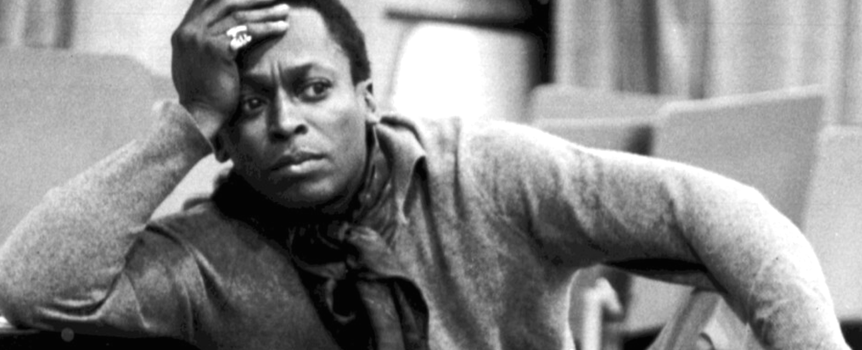 Miles Davis w szarym ubraniu z papierosem, zamyślony, siedzi na krześle w pomieszczeniu.