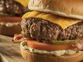 Zdjęcie przedstawia dwa soczyste burgery z serem, pomidorem, boczkiem i marynowanym ogórkiem.