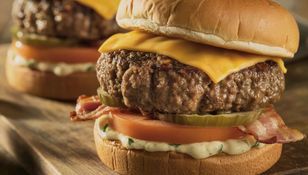 Zdjęcie przedstawia dwa soczyste burgery z serem, pomidorem, boczkiem i marynowanym ogórkiem.