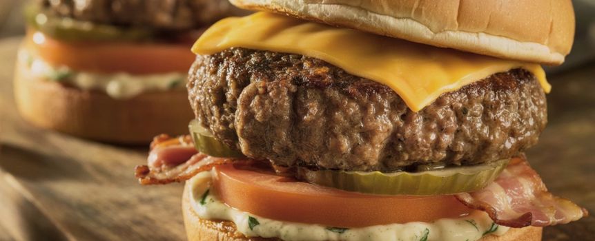 Zdjęcie przedstawia dwa soczyste burgery z serem, pomidorem, boczkiem i marynowanym ogórkiem.