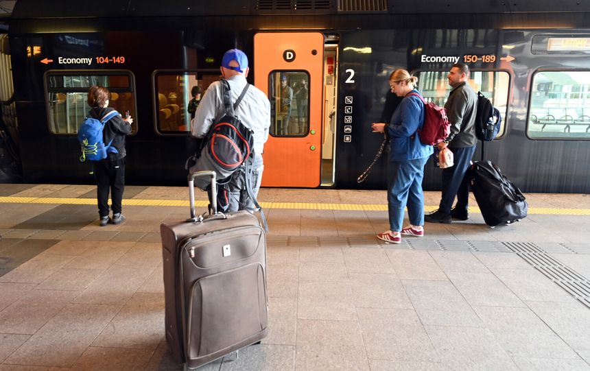 Grupa osób czeka przy pociągu z bagażami na stacji. W tle widoczne informacje o trasach.
