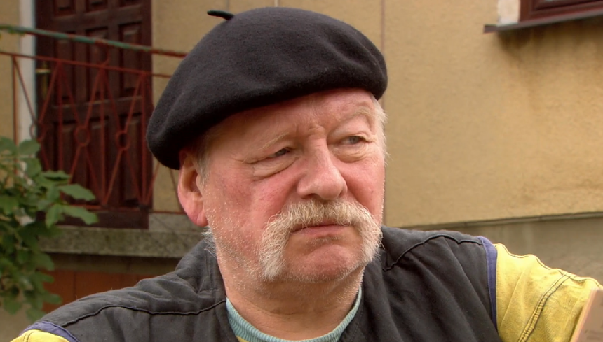 Mężczyzna w czapce beretowej z wąsami, siedzi w plenerze, z tłem ściany budynku.