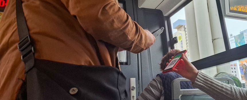 Osoby w autobusie, jedna skanuje bilet telefonem, druga trzyma telefon z kodem QR.