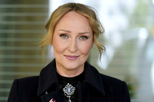 Anita Lipnicka w czarnym płaszczu, z uśmiechem, stojąca na tle rozmytej przestrzeni.