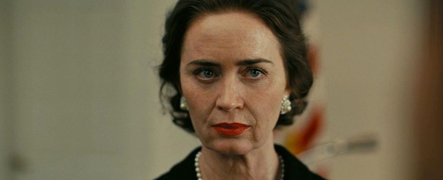 Na zdjęciu kobieta w eleganckiej czarnej sukience i perłowej biżuterii, z zamyśloną miną.