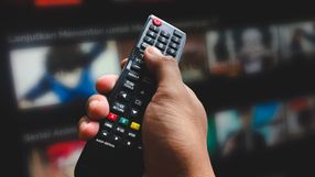 Ręka trzymająca pilot do telewizora, w tle zarysy ekranów z serialami animowanymi.