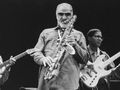 Muzycy jazzowi na scenie; mężczyzna gra na saksofonie, w tle gitarzysta i basista.