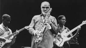 Muzycy jazzowi na scenie; mężczyzna gra na saksofonie, w tle gitarzysta i basista.