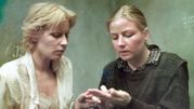 Dwie kobiety w trudnej rozmowie, jedna z nich dotyka dłoni drugiej. Tło w odcieniach szarości.