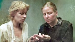 Dwie kobiety w trudnej rozmowie, jedna z nich dotyka dłoni drugiej. Tło w odcieniach szarości.