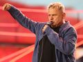 Mężczyzna z mikrofonem, w niebieskiej koszuli, wyraża emocje na scenie z czerwonym tłem.