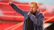Mężczyzna z mikrofonem, w niebieskiej koszuli, wyraża emocje na scenie z czerwonym tłem.