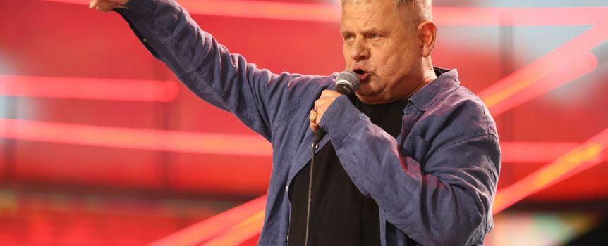 Mężczyzna z mikrofonem, w niebieskiej koszuli, wyraża emocje na scenie z czerwonym tłem.