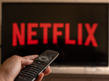 Pilot telewizyjny trzymany w dłoni, a w tle logo Netflix na ekranie telewizora.