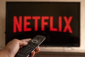 Pilot telewizyjny trzymany w dłoni, a w tle logo Netflix na ekranie telewizora.