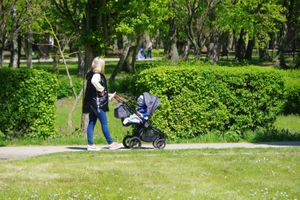 Kobieta z wózkiem spacerowym przechadza się parkiem wśród zieleni drzew i krzewów.