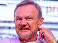 Cezary Pazura w jasnej marynarce, uśmiecha się, siedząc na tle kolorowych świateł.