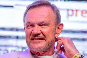 Cezary Pazura w jasnej marynarce, uśmiecha się, siedząc na tle kolorowych świateł.