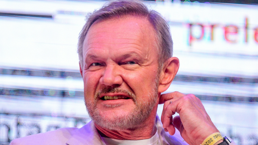 Cezary Pazura w jasnej marynarce, uśmiecha się, siedząc na tle kolorowych świateł.