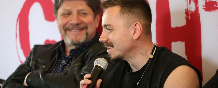 Na zdjęciu mężczyzna z mikrofonem w ręku, obok uśmiechający się mężczyzna w skórzanej kurtce.