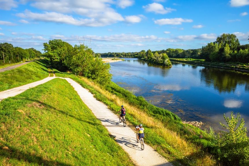 Dwóch rowerzystów na ścieżce wzdłuż rzeki, zieleń wokół, jasne niebo z chmurami.