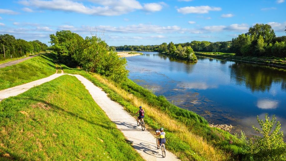Dwóch rowerzystów na ścieżce wzdłuż rzeki, zieleń wokół, jasne niebo z chmurami.