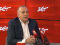 Mężczyzna w garniturze siedzi przy stole, mówi do mikrofonów w studio radiowym. Tło w kolorze czerwonym.