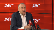 Mężczyzna w garniturze siedzi przy stole, mówi do mikrofonów w studio radiowym. Tło w kolorze czerwonym.