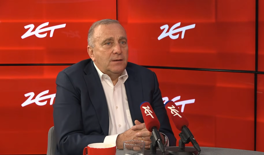 Mężczyzna w garniturze siedzi przy stole, mówi do mikrofonów w studio radiowym. Tło w kolorze czerwonym.
